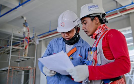 Two engineers in safety helmets reviewing construction plans at a worksite.